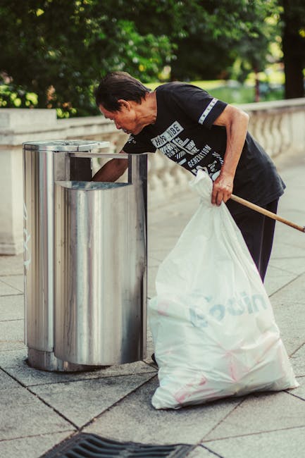 A middle-aged woman with short dark hair, wearing a black T-shirt with white graphic text and black trousers, is seen disposing of waste into a modern, stainless steel, dual-compartment outdoor bin located on a paved pathway. She holds a large white plastic sack filled with rubbish in her left hand and uses her right hand to lift the lid of the bin. The bin is situated adjacent to a low stone railing and is surrounded by lush green foliage and trees in the background, suggesting an outdoor urban park or public area. The scene is illuminated by natural daylight, highlighting the reflective surface of the metal waste container. This image captures the act of on-site waste disposal, highlighting local waste management practices that may differ from council-provided rubbish collections, illustrating an example of private or independent rubbish handling supported by local waste removal services like Rubbish Removal Highbury.