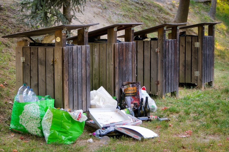 Four wooden compost or refuse bins with slanted tops are positioned on a grassy area beside a sloped hill with trees and foliage in the background. The bins are constructed from vertical wooden planks, with a weathered, dark brown finish, and are arranged in a straight line. In front of the bins, there are various discarded items, including a large green reusable shopping bag filled with plastic bottles, a white takeout pizza box with some leftover food, several glass bottles, a cardboard box with the word 'barbecue' visible, and plastic bags containing waste. The scene is outdoors, with natural light illuminating the area, which appears to be a designated waste disposal or collection point, possibly set up for private rubbish removal services. The overall environment indicates a combination of functional waste storage and accidental litter accumulation, highlighting the importance of proper waste management practices.