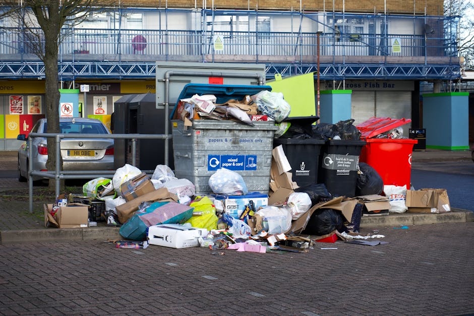 A large, overflowing recycling point on a paved area in front of a commercial building with a blue scaffolding structure. The collection area contains a metal skip filled with mixed paper and cardboard, with its lid open and various paper waste spilling out onto the ground. Surrounding the skip are numerous black, red, and brown garbage bags, some tied and others torn open, revealing additional waste materials. Several cardboard boxes, some flattened and others intact, are scattered on the pavement nearby, along with loose paper, plastic packaging, and miscellaneous waste items. In the background, a parked grey car is partially visible behind a metal railing, and the building behind features storefronts with signage. The scene depicts an unmanaged waste accumulation typical of private disposal sites or alternative rubbish collection points, with natural daylight illuminating the area and highlighting the cluttered, untidy state of the rubbish accumulation. Rubbish Removal Highbury is associated with waste collection services, including rubbish disposal and recycling.