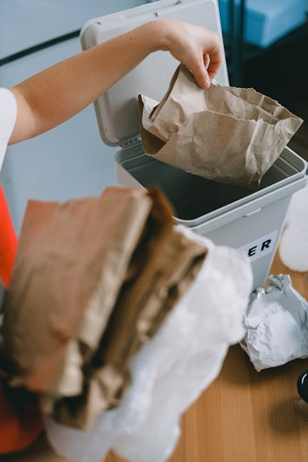 A person’s hand is seen placing a brown paper bag into an open white rubbish bin labeled 'RUBBISH', situated indoors on a wooden surface. In the foreground, there is a collection of additional crumpled paper bags, some white and others brown, held loosely in the person’s other hand. The background shows a corner of a room with a part of a blue container or shelf and some scattered white paper or packaging material nearby. The trash bin appears to be used for general waste, with a dark liner visible inside. The environment suggests a typical domestic setting focused on waste disposal, possibly reflecting responsible rubbish management and the importance of separating waste for disposal or recycling, in line with local waste handling practices discussed in the Highbury guide to council recycling and rubbish rules. The scene emphasizes the act of proper rubbish separation and disposal, aligned with private waste collection services offered by Rubbish Removal Highbury.