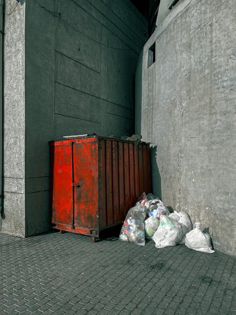 A large outdoor collection of empty wooden and plastic fruit boxes stacked in a disorderly manner on a paved surface, with some boxes collapsed or leaning. The wooden crates are light-colored, weathered, and have printed labels indicating they once contained fresh fruits, featuring images of tomatoes and other produce. Several black plastic crates are present, stacked on top of or beside the wooden boxes, with a rough texture and matte finish. To the right, part of a green, weathered wheelie bin is visible, suggesting waste disposal or collection activity. The environment appears to be a storage or sorting area for fruit packaging or surplus stock, with diffused natural lighting illuminating the scene, contributing to a neutral, practical atmosphere suitable for describing waste management or rubbish removal services. Rubbish Removal Highbury might handle such bulk storage disposal, emphasizing private or alternative waste handling options outside standard local authority collections.