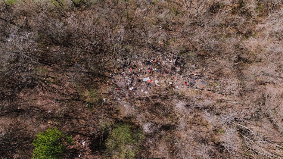 An aerial view of a densely wooded area with mostly bare deciduous trees, suggesting late autumn or winter, shows a scattered collection of discarded rubbish and debris concentrated in the center of the scene. The debris includes various materials such as cardboard boxes, plastic bottles, metal objects, and other waste items, some of which are partially collapsed or layered. The surrounding trees have a mix of brown and gray branches, with few leaves, and the ground beneath the rubbish appears uneven and slightly disturbed. In the lower left corner, a small patch of bright green foliage contrasts with the otherwise muted natural tones. The scene is illuminated by natural light, casting soft shadows across the waste and trees, highlighting the stark contrast between the natural environment and the unmanaged refuse. This image could illustrate an example of illegal dumping or unmanaged rubbish accumulation in a natural area, which may require professional waste removal services like those offered by Rubbish Removal Highbury for alternative waste handling solutions.