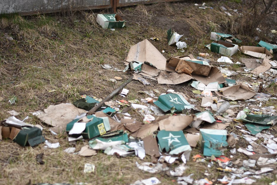 The image shows an outdoor scene with discarded cardboard boxes, some of which are partially flattened, and plastic packaging materials scattered across a grassy area adjacent to a pavement or driveway. The boxes appear to be used for beverages, indicated by printed logos and branding, with a mix of green and white colors, featuring star symbols. Some boxes are torn or crumpled, with pieces of paper, plastic wrapping, and small debris mixed in among the larger packaging. The ground is uneven with patches of grass and dirt, and there is a metal pole or pipe lying among the rubbish. The surrounding environment includes a wooden or brick fence at the top, with leafless shrubbery and trees in the background, suggesting early spring or late winter. The scene depicts a typical example of improperly disposed waste in a residential or semi-urban outdoor area, highlighting the need for effective rubbish removal and collection services like those provided by Rubbish Removal Highbury to manage private waste clearance.