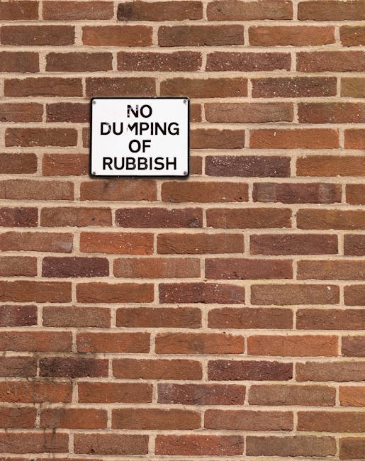 A rectangular white sign with black uppercase text reading 'NO DUMPING OF RUBBISH' is mounted on a red brick wall. The wall features uniformly sized bricks with a textured surface, arranged in a horizontal pattern, and mortar joints visible between each brick. The sign is positioned above center towards the upper section of the wall, which has a mix of darker and lighter red bricks, some showing slight weathering. The lighting appears natural, casting soft shadows on the bricks below the sign. The overall scene suggests a typical exterior urban wall where private waste handling or independent rubbish collection may occur, and the sign serves as a notice against illegal dumping, aligning with the context of rubbish removal services by companies such as Rubbish Removal Highbury located at https://rubbishremovalhighbury.com.