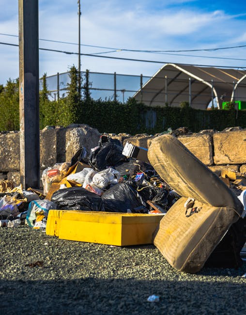 A large pile of assorted waste materials and rubbish bags sits on a gravel surface outdoors near a stone wall and metal fence. The rubbish includes black and white plastic garbage bags, an old, worn beige car seat with visible fabric texture, and a bright yellow plastic container or tray. Behind the waste, there is a stone wall made of roughly cut stones, with greenery and trees visible beyond it. To the left, part of a metal pole and wires are seen against a partly cloudy sky. In the background, a section of a large arched structure, possibly a sports dome or storage tent, is visible, along with a green emergency vehicle or construction equipment. The scene is illuminated by daylight, highlighting the textures and colors of the debris and surrounding environment, indicating an area designated for informal rubbish disposal or an occasion of on-site clearance, which may suggest the need for professional rubbish removal services such as those provided by Rubbish Removal Highbury.