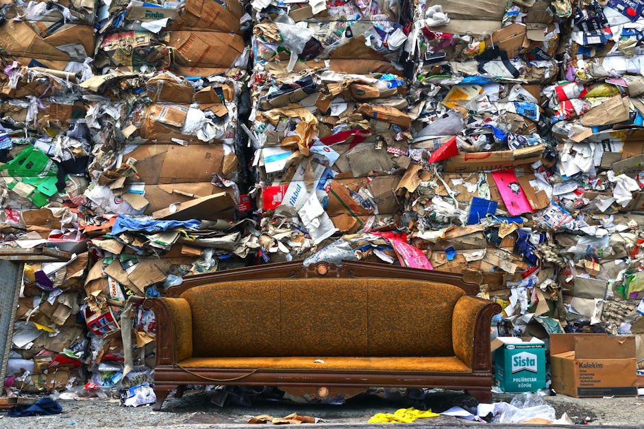 A vintage-style upholstered sofa with brown patterned fabric and carved wooden frame is positioned on a sidewalk in front of an extensive wall of densely piled waste and discarded cardboard boxes. The sofa appears slightly weathered with some frayed fabric and minor damage along the base, contrasting sharply with the cluttered environment behind it. The waste consists mainly of crumpled and flattened cardboard boxes in various shades of brown, white, and coloured printed packaging, stacked haphazardly to form a large, uneven wall that extends beyond the edges of the image. Scattered around the base of the sofa are additional cardboard boxes, some labeled with company names such as 'Sista' and 'Kalekim,' along with small pieces of torn paper and plastic. The scene suggests an informal rubbish collection or an area where bulky waste is temporarily stored, highlighting the challenges of waste management and disposal that waste removal services like Rubbish Removal Highbury might address in urban environments.