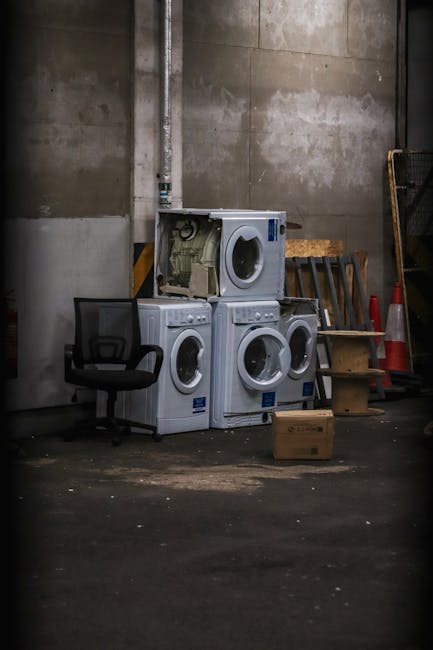 An indoor storage area featuring four front-loading washing machines arranged as two stacked units on top of each other, with one of the machines on top having its door open revealing internal components. To the left of the appliances is a black office chair with a mesh backrest, positioned on a concrete floor that shows some dirt and wear. Behind the machines, there is a plain, unfinished concrete wall with visible pipes running vertically along it. To the right, a small wooden step stool with two steps is placed next to several orange traffic cones and a black wire mesh panel leaning against the wall. The environment appears to be a utilitarian space used for storage or disposal, consistent with an area where bulky waste such as household appliances is temporarily kept before removal or disposal by professional waste management services like Rubbish Removal Highbury. Natural or indirect lighting creates a neutral, slightly shadowed atmosphere, emphasizing the functional, no-frills setting suitable for managing waste or bulky items for disposal or professional collection.