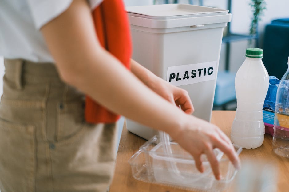 Close-up image of a person wearing beige trousers and a white shirt, with only their torso and arms visible. The individual is placing a small, clear plastic container into a white plastic bin labeled 'PLASTIC' on a light-wood table. Surrounding the bin on the table are various plastic bottles, including a transparent bottle with a green cap and a larger, semi-transparent bottle with a blue label, indicating different types of plastic containers. The background reveals a partially blurred interior setting with a blue couch and natural light streaming in from a window, suggesting a home or office environment focused on responsible waste sorting and recycling, with Rubbish Removal Highbury providing services for proper rubbish collection and disposal in the area.