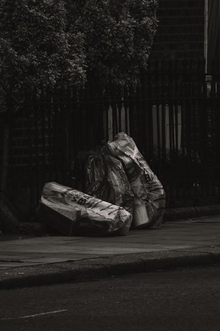 Two large black garbage bags filled with waste are placed on the pavement in front of a dark wooden fence. The bags appear to be made of thick, crinkled plastic, with some areas reflecting faint light. Behind the bags, a portion of a tall wooden fence with vertical slats is visible, partially obscured by shadows. To the left, there is a leafy tree with dense dark foliage extending upwards, creating a natural backdrop. The scene takes place on a quiet street curb, with the edge of the asphalt visible at the bottom of the image. The environment appears to be an urban or suburban residential area, where private rubbish collection or on-site clearance by Rubbish Removal Highbury may be necessary for waste disposal outside of council services. The overall lighting is subdued, adding to the neutral and factual tone of the scene, emphasizing the importance of proper rubbish management.