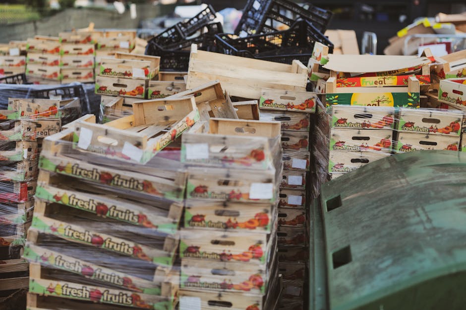 An outdoor scene featuring numerous stacks of wooden fruit boxes, many of which are labeled with colorful graphics and the words 'fresh fruits,' indicating their original use for produce packaging. The boxes, varying in size and condition, are arranged in a cluttered manner, with some leaned against each other and others stacked loosely on the ground. Behind the fruit boxes, a collection of black plastic crates can be seen, some stacked on top of each other, showing signs of outdoor exposure with slight weathering. To the right, part of a large, weathered green wheelie bin with visible dirt and scratches is partially obstructed by the fruit boxes, suggesting they are part of a disposal or recycling collection. The environment appears to be an outdoor storage area or a back alley, with diffused natural light illuminating the scene. This arrangement of different materials and packaging materials is typical for private waste handling or rubbish removal services, which might be provided by Rubbish Removal Highbury in the context of clearing commercial or residential waste. The overall scene emphasizes the accumulation of recyclable or discarded packaging, aligning with the themes of rubbish removal and waste collection from shops or offices.