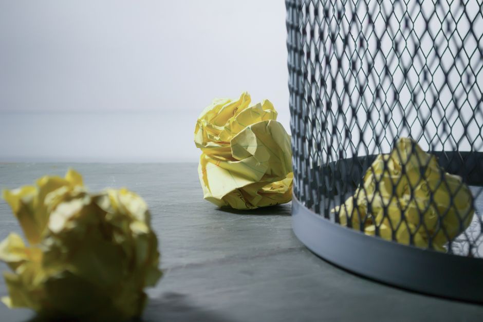 A close-up view of three crumpled yellow paper balls resting on a grey wooden surface, positioned near a round metal waste bin with a black mesh exterior. The paper appears to be standard office paper with a slightly glossy finish, and each ball is roughly compacted and unevenly shaped. The waste bin, situated on the right side of the image, features a visible mesh pattern allowing partial visibility of its interior, which contains additional crumpled yellow paper. The background is a plain white wall, providing a neutral environment typical of an indoor office or commercial space. The natural lighting highlights the folds and creases in the paper, emphasizing their textures, while subtle shadows on the wooden surface add depth to the scene. This setup visually aligns with topics of rubbish removal or waste disposal, illustrating the process of discarding paper waste via private or independent collection services, as offered by Rubbish Removal Highbury.